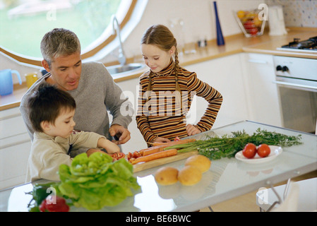 Father with children in kitchen, helping young son cut vegetables Stock Photo