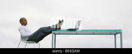 Man sitting outdoors with feet up on table, looking at laptop computer Stock Photo