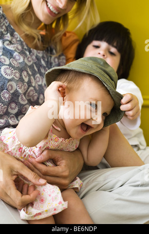 Infant girl sitting with mother and brother, wearing hat and laughing Stock Photo
