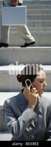 Businesswoman using cell phone outdoors, person walking on steps in background Stock Photo