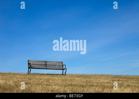 An empty park bench made of wood on dry grass is outlined against a blue sky with light clouds. Stock Photo