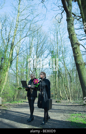 Two businesswomen walking together along forest path, one using laptop, both carrying artificial flowers Stock Photo