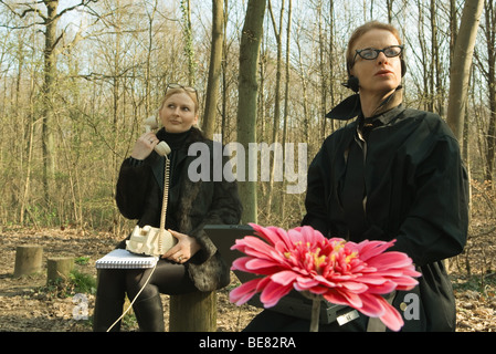 Woman sitting in wooded area using landline telephone, companion sitting nearby, looking away Stock Photo