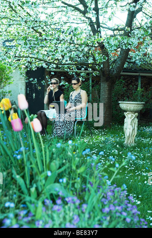 Two women sitting in garden using laptop computer and landline phone Stock Photo
