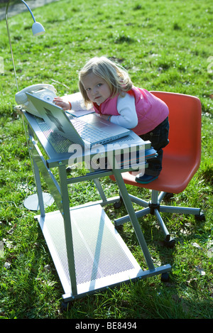 Little girl standing on office chair by desk in field, playing with laptop computer, looking at camera Stock Photo