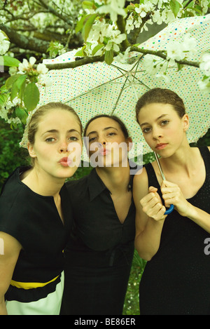 Three female friends standing under umbrella together, two blowing kisses at camera Stock Photo