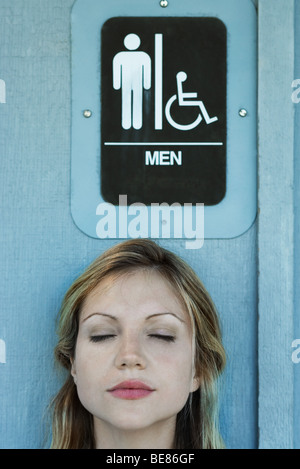 Woman in front of men's restroom sign, eyes closed Stock Photo