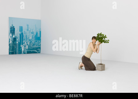 Woman kneeling, hugging potted tree, cityscape in background Stock Photo