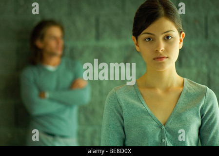 Young woman looking at camera, man in background leaning against wall with arms folded Stock Photo