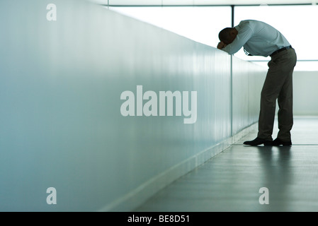Man standing in corridor, bending over with head resting on arms, side view Stock Photo