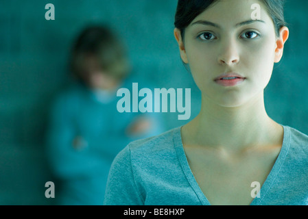 Young woman looking at camera, man in background leaning against wall with arms folded and head lowered Stock Photo