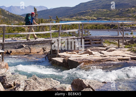 Young woman crossing creek, wooden bridge, Deep Lake behind, hiking, backpacking, hiker with backpack, historic Chilkoot Trail, Stock Photo