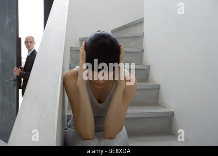 Woman sitting on stairs with hands over ears, man leaving in background Stock Photo