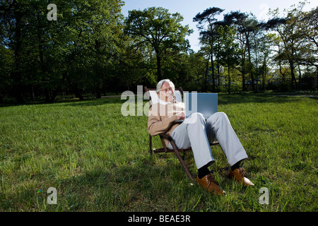 A senior man sitting in a lounge chair using a laptop, outdoors Stock Photo