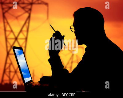 man with laptop computer looking out window at power poles silhouetted at sunset Stock Photo