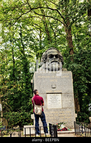 A man in a maroon shirt stands gazing up in front of the grave of Karl Marx in Highgate Cemetery London England. Stock Photo