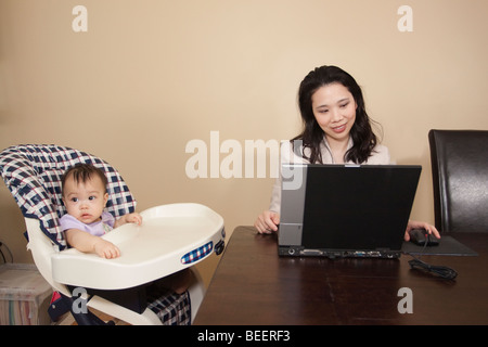 Chinese mother working on laptop next to baby in high chair Stock Photo