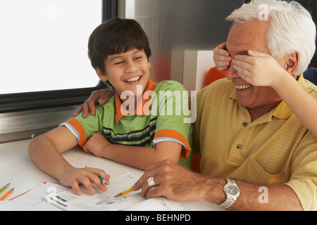 Senior man helping his grandson in his homework and a child's hands covering his eyes from behind Stock Photo