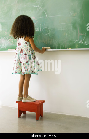 Little girl standing on stool in front of blackboard, rear view Stock Photo