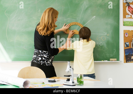 Teacher helping boy draw angle on blackboard using protractor, rear view Stock Photo