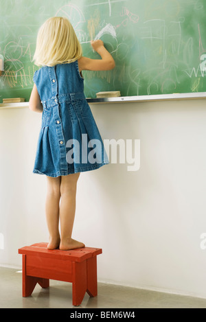 Little girl standing on stool, scribbling on blackboard, rear view Stock Photo