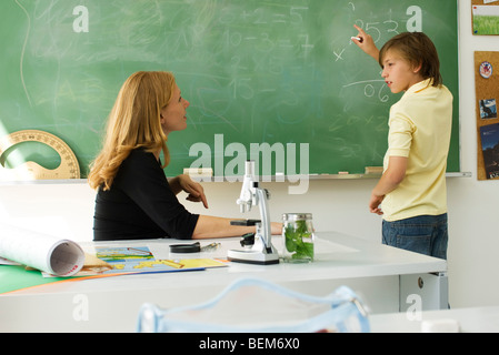 Boy solving math problem on blackboard, looking at teacher for assistance Stock Photo