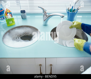 Close-up of hands wearing yellow rubber gloves washing dishes in double kitchen sink with stainless steel tap pouring water Stock Photo