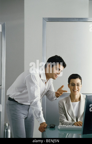 Office scene, man leaning on edge of desk speaking to female colleague, both looking at her computer screen Stock Photo