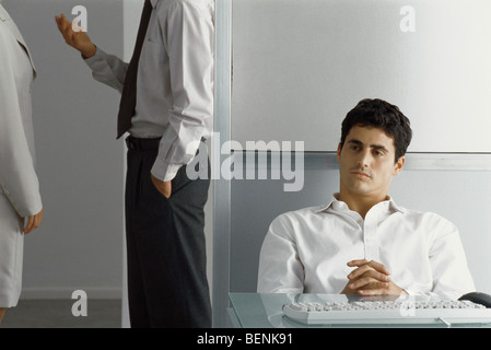 Man sitting at desk with hands clasped while colleagues talk nearby Stock Photo