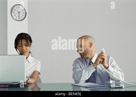 Office worker sitting at desk holding phone, covering receiver and speaking to female co-worker Stock Photo
