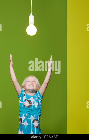 Little girl with arms raised, reaching for light bulb suspended overhead Stock Photo