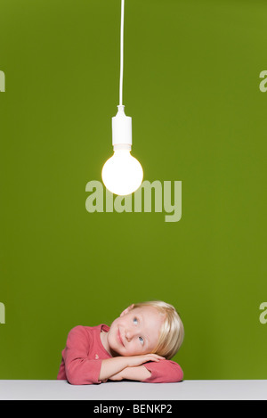 Little girl resting head on arms contemplating illuminated light bulb suspended overhead Stock Photo