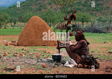 Himba woman cooking in front of traditional mud hut, Kaokoland / Kaokoveld, Kunene Region, Northern Namibia, South Africa Stock Photo