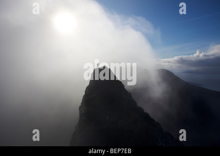 A lone climber stands on a mountain summit in the Black Cuillins, with a mist shrouded sun behind him, Isle of Skye, Scotland Stock Photo