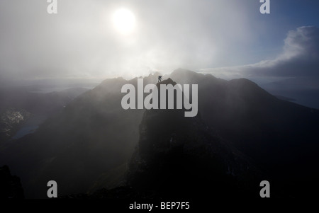 A lone climber stands on a mountain summit in the Black Cuillins, with a mist shrouded sun behind him, Isle of Skye, Scotland Stock Photo