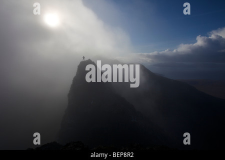 A lone climber stands on a mountain summit in the Black Cuillins, with a mist shrouded sun behind him. Isle of Skye, Scotland Stock Photo