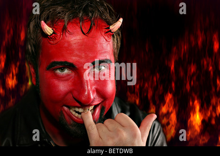 Devilish man looking naughty and coy while biting on his pinky, with flames of fire in the background. Stock Photo