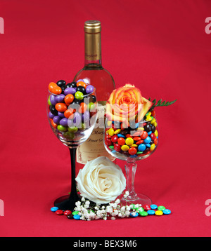 A bottle of wine two wineglasses filled with M&M candy and two beautiful roses. Shot against a red background. A holiday shot. Stock Photo