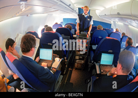 Airline passengers using computer laptops in business class with flight attendant offering duty free items from cart Stock Photo