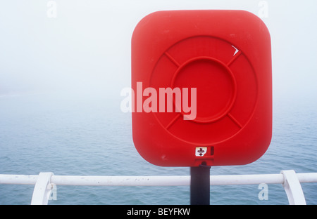 Detail of red life-saving equipment on pole by white railings with calm sea in fog beyond Stock Photo