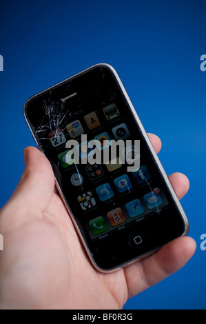 An damaged Apple iPhone 3G being held in front of a blue background.  The main display glass has been cracked. Stock Photo