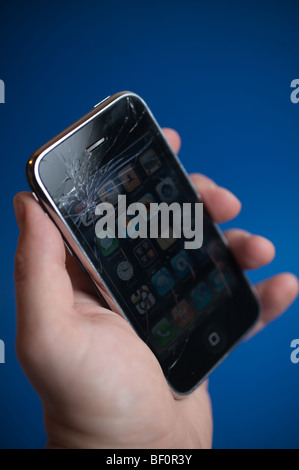 An damaged Apple iPhone 3G being held in front of a blue background.  The main display glass has been cracked. Stock Photo