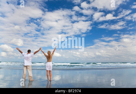 A young man and woman celebrating arms raised and holding hands as a romantic couple on a beach with a bright blue sky Stock Photo