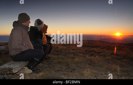 Three hikers dressed in warm clothing watching the sun rise over the horizon, Cathedral Peak, Drakensberg Ukahlamba National Par Stock Photo