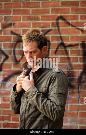 A 30 year old man alone wearing scruffy jacket standing against brick wall looking depressed sad alone lonely Stock Photo