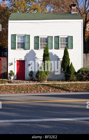 white painted saltbox house with asymmetric windows green shutters bright red door & ornamental cedars in Montgomery New York Stock Photo