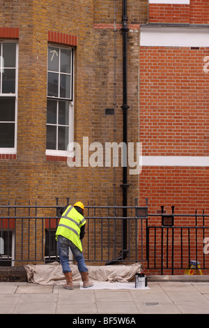 Builder worker maintence man painting iron railing outside London apartment blocks wearing high viz jacket safety helmet Stock Photo