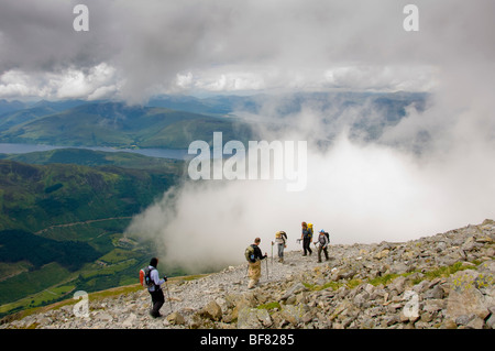 Walkers using trekking poles on the rocky path, above the clouds on Ben Nevis with Loch Eil in the distance. Scotland Stock Photo