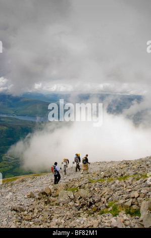 Walkers using trekking poles on the rocky path, above the clouds on Ben Nevis with Loch Eil in the distance. Scotland Stock Photo