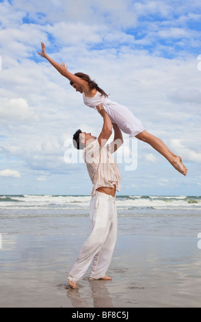 A young man holding his girlfriend above is head as she jumps in celebration on a beach Stock Photo